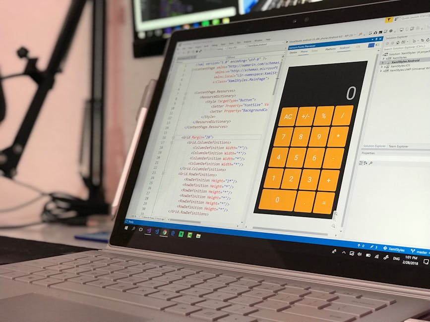 A laptop seen from an angled perspective displaying a code editor with HTML and CSS code on the screen, alongside a large orange calculator application open beside it. The workspace is set up on a des