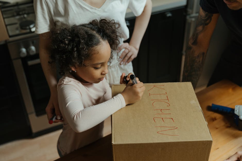 A man with curly black hair and a beard is inside a well-lit room with white walls and wooden flooring, carrying a medium-sized cardboard box with red tape around it. He is dressed in a blue T-shirt w